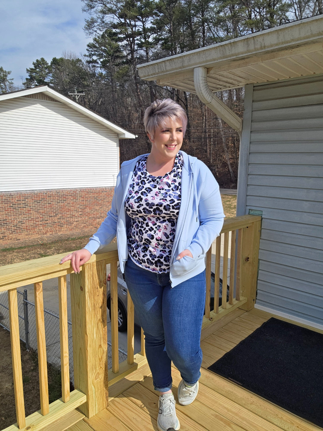 Woman wearing LuLaRoe Essential Zip Hoodie in Solid Cloud Blue fleece with zip front, drawstring hood, and kangaroo pocket paired with denim jeans on a wooden deck with a house and trees in the background