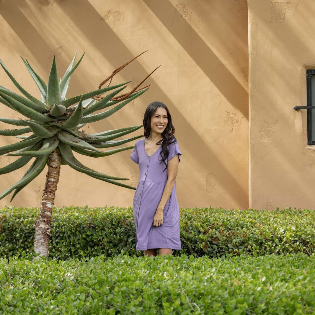 Woman wearing LuLaRoe Heidi babydoll dress in solid dusty lavender purple with button front, short sleeves, and flowy fit standing outdoors near greenery and a stucco wall