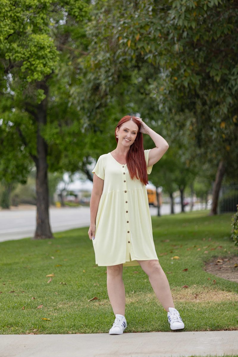 Woman wearing LuLaRoe Heidi babydoll dress in solid butter yellow with button front, short sleeves, flowy fit, and pockets standing outdoors on grass with trees in the background
