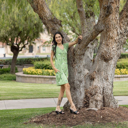 Woman wearing LuLaRoe Heidi babydoll dress in green floral print with white flowers, button front, short sleeves, and flowy fit standing outdoors by a tree