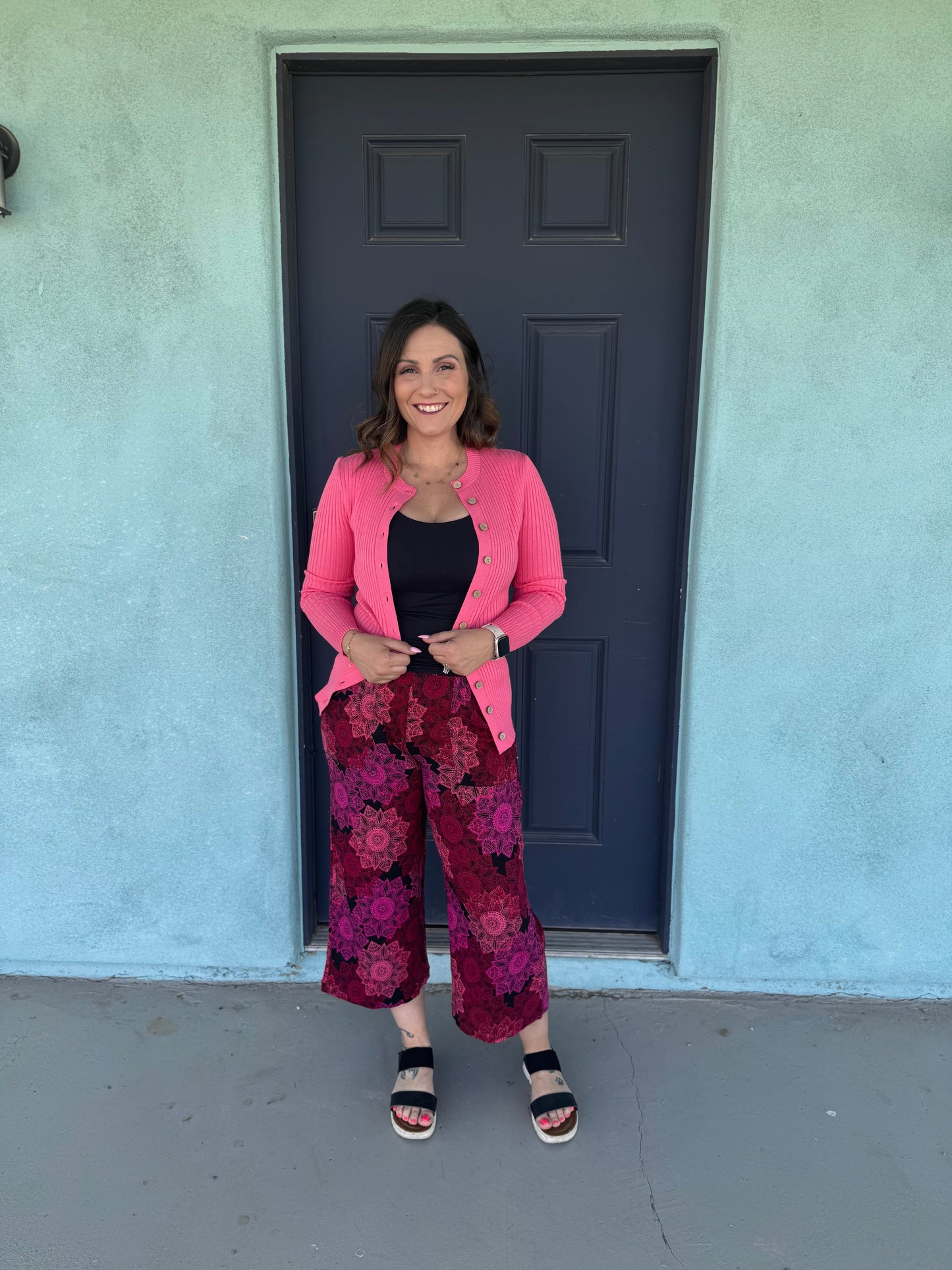 Woman standing in front of a blue door wearing LuLaRoe Justine wide-leg pants in red and black mandala floral print, featuring elastic waistband and cropped relaxed fit, styled with black top and pink cardigan for a bold outfit.