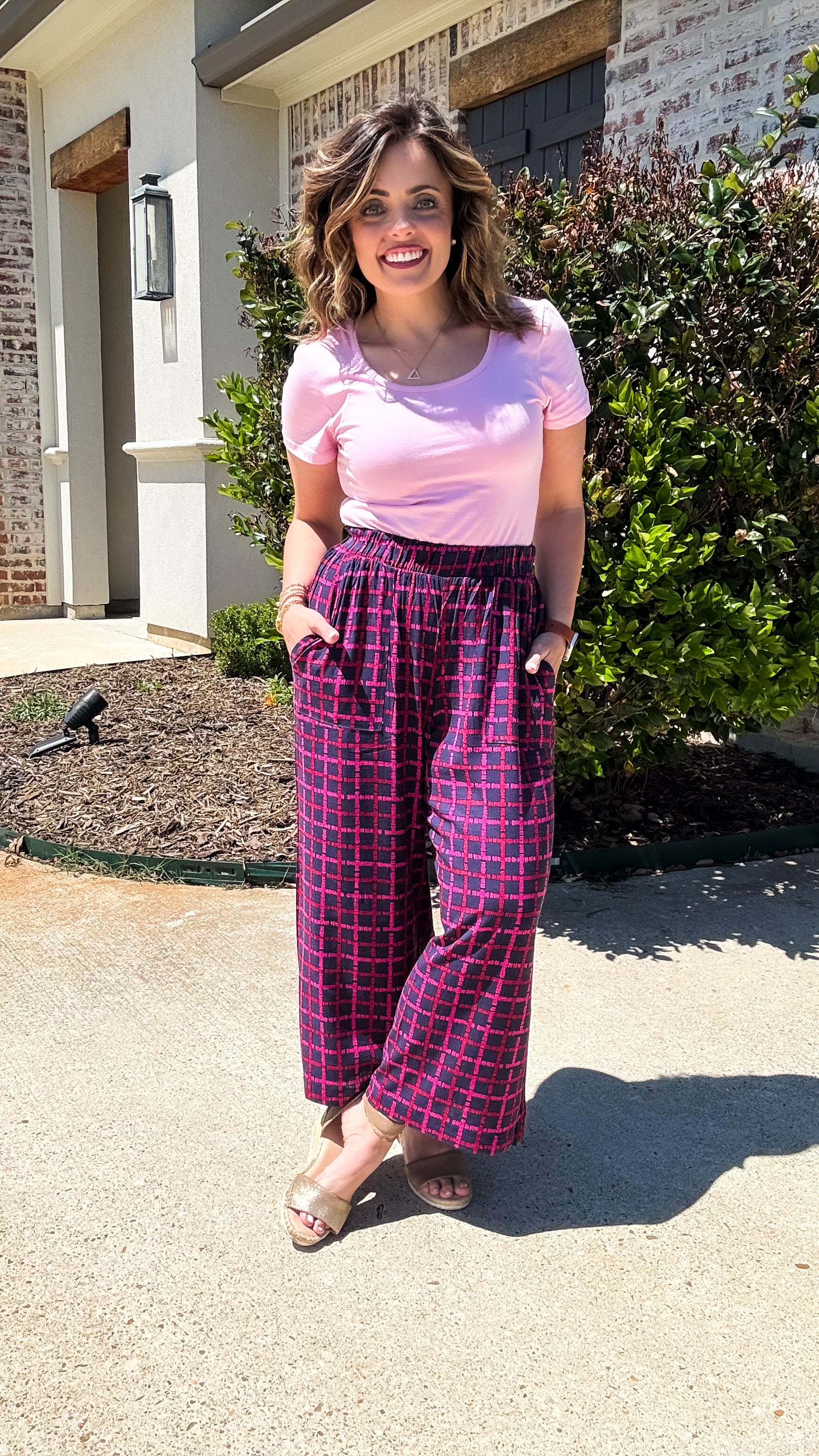 LuLaRoe Justine wide-leg pants in plum with raspberry pink grid pattern, featuring elastic waistband, relaxed fit, and cropped wide-leg silhouette worn styled with a pink top outdoors standing outside a house.