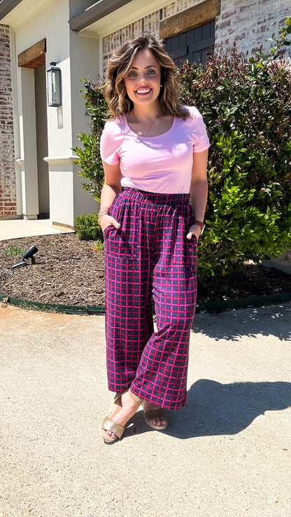 LuLaRoe Justine wide-leg pants in plum with raspberry pink grid pattern, featuring elastic waistband, relaxed fit, and cropped wide-leg silhouette worn styled with a pink top outdoors standing outside a house.