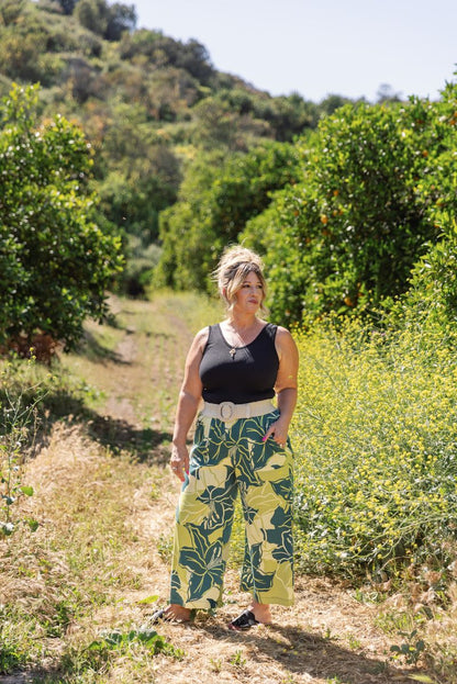 LuLaRoe Justine wide-leg pants in green and yellow tropical leaf print, featuring elastic waistband and cropped relaxed fit, styled outdoors with a black tank for a resort-inspired outfit standing on a dirt path surrounded by greenery
