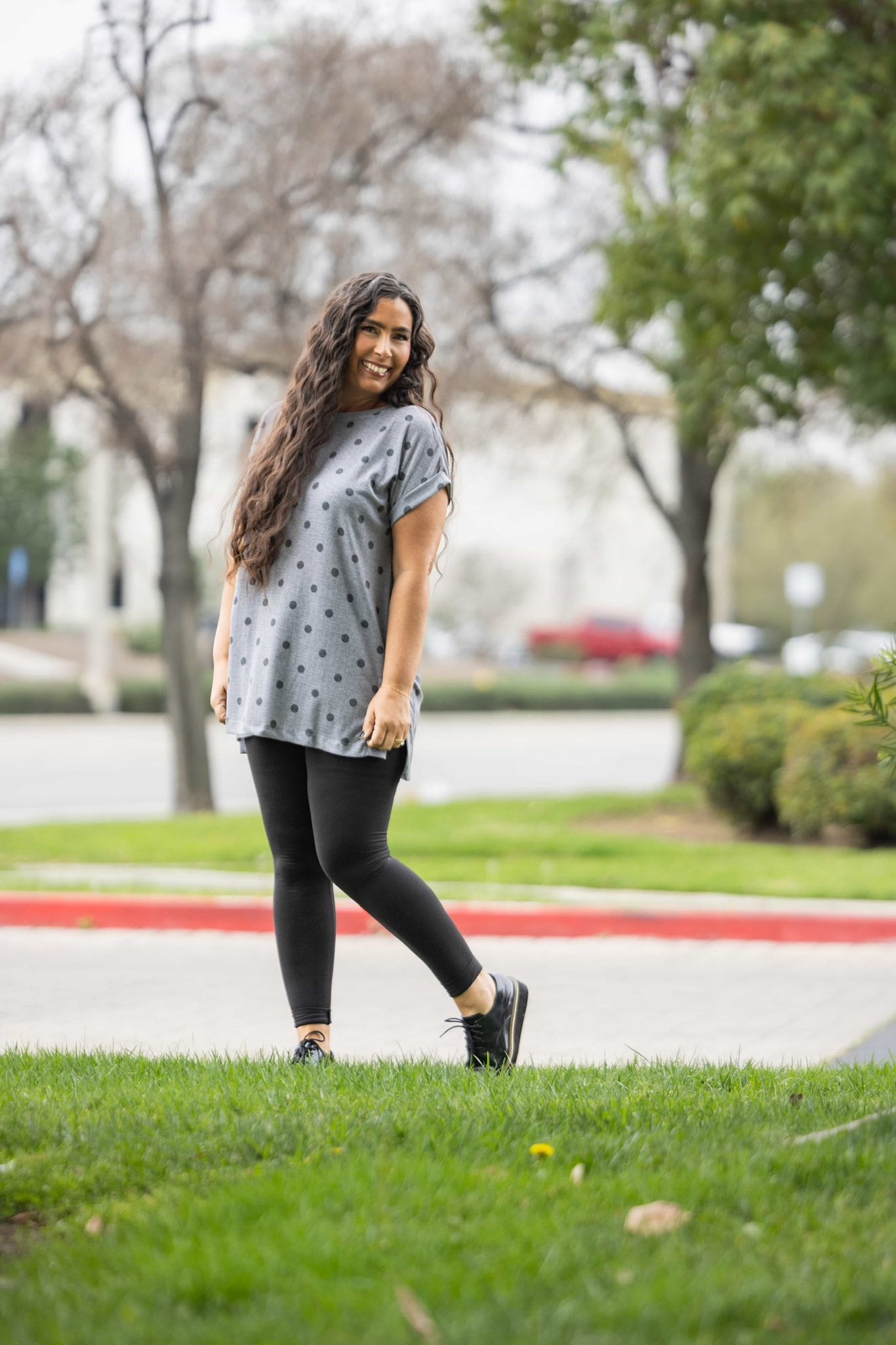 Woman wearing LuLaRoe Leah top in heather gray with charcoal polka dot print, relaxed fit women’s boutique polka dot top, casual everyday layering piece outdoors on a grassy area with trees and a road in the background
