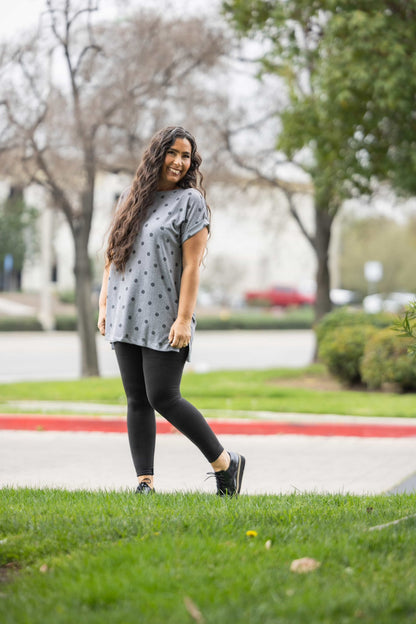Woman wearing LuLaRoe Leah top in heather gray with charcoal polka dot print, relaxed fit women’s boutique polka dot top, casual everyday layering piece outdoors on a grassy area with trees and a road in the background