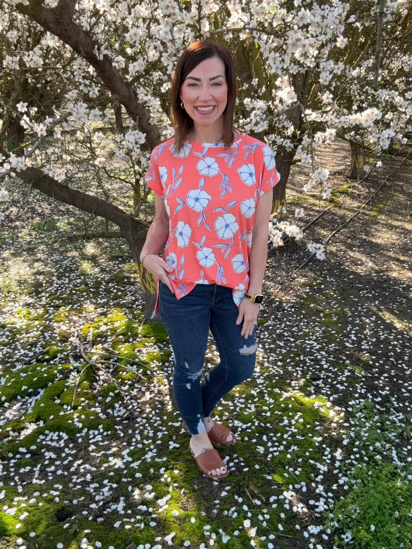 LuLaRoe Leah top in coral pink with white and blue floral print, relaxed fit women’s boutique floral top for spring and summer on a model in a floral top standing under a cherry blossom tree