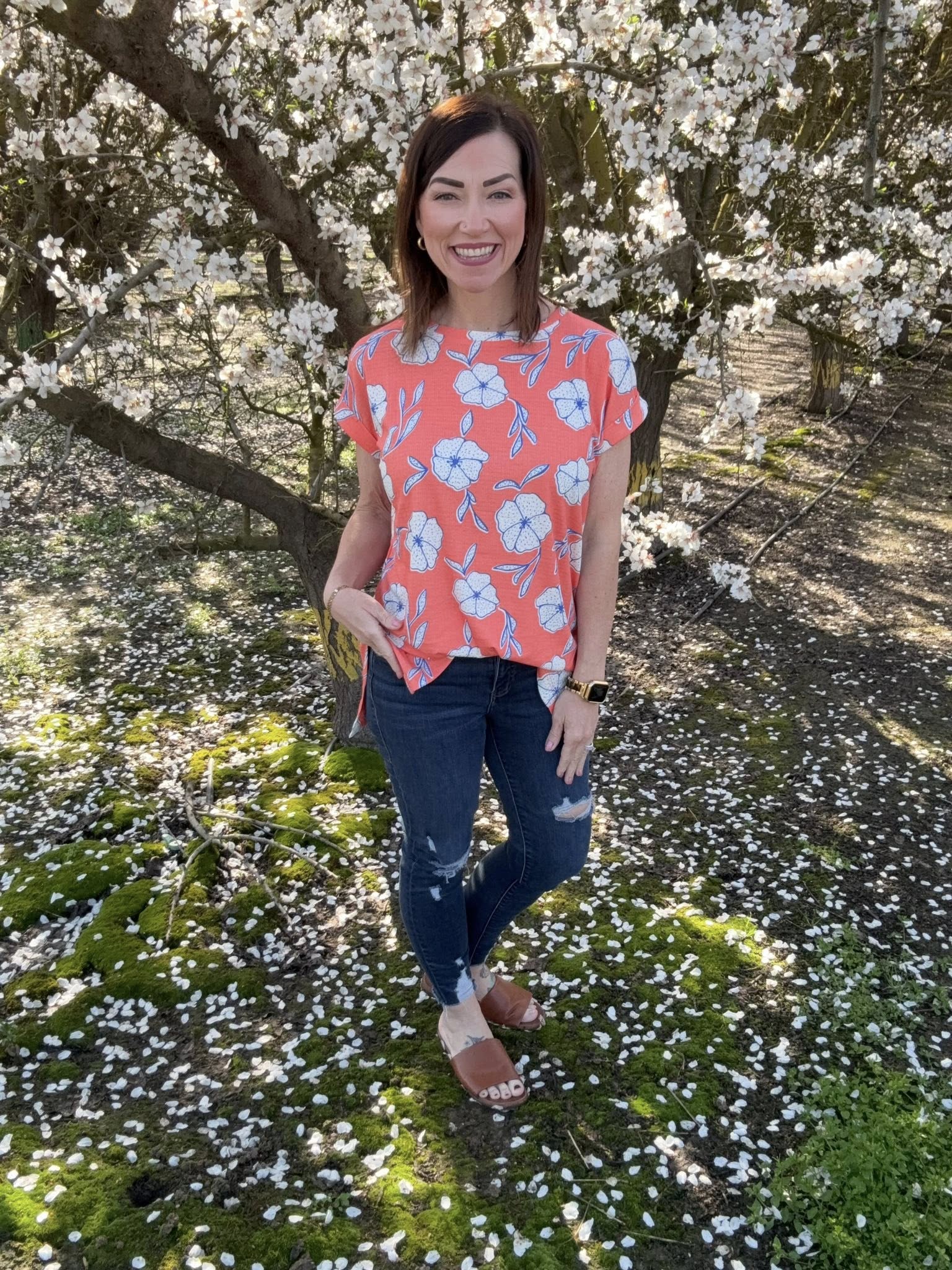LuLaRoe Leah top in coral pink with white and blue floral print, relaxed fit women’s boutique floral top for spring and summer on a model in a floral top standing under a cherry blossom tree