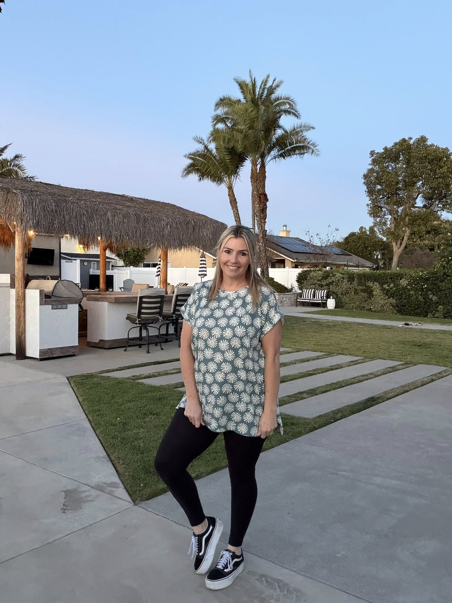 LuLaRoe Leah top in sage green with white daisy burst floral print, relaxed fit women’s boutique casual top on a woman standing in a backyard with outdoor furniture and palm trees.