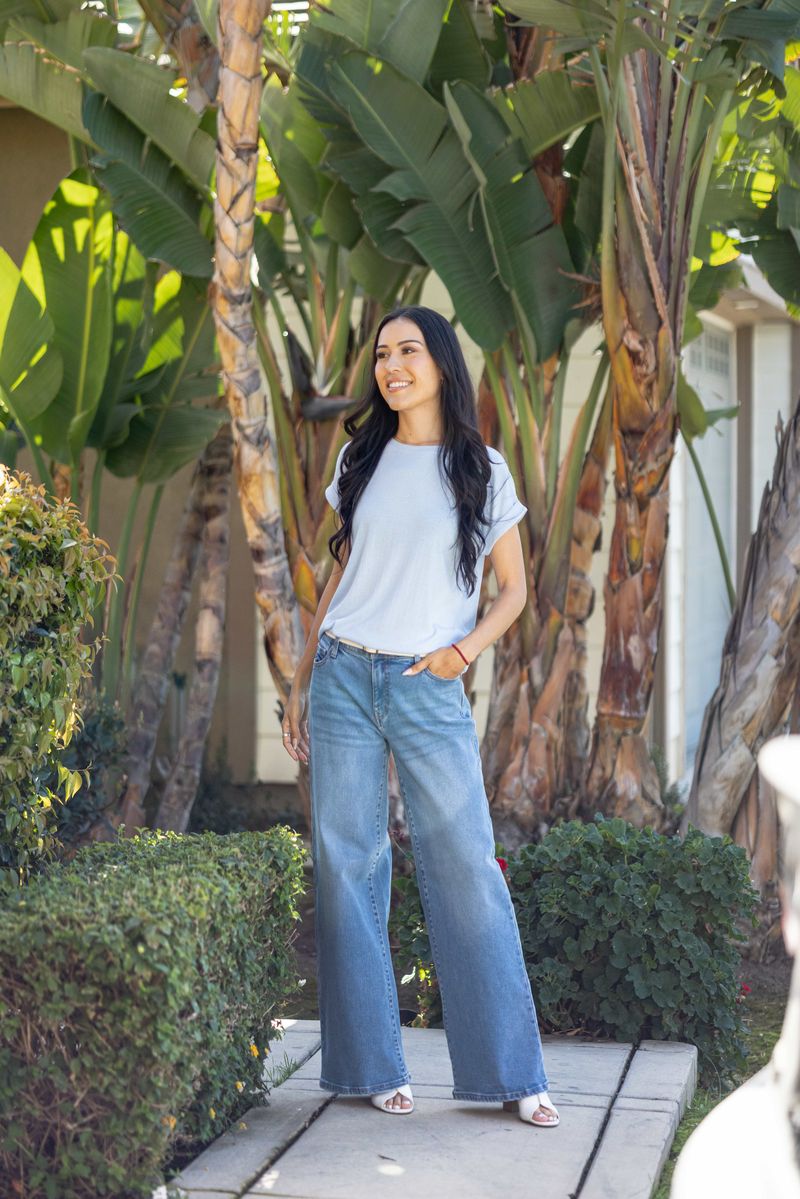 LuLaRoe Leah top in solid sky mist blue, relaxed fit women’s light blue boutique top with soft drape fabric, versatile everyday layering piece on a woman standing outdoors with palm trees and greenery in the background