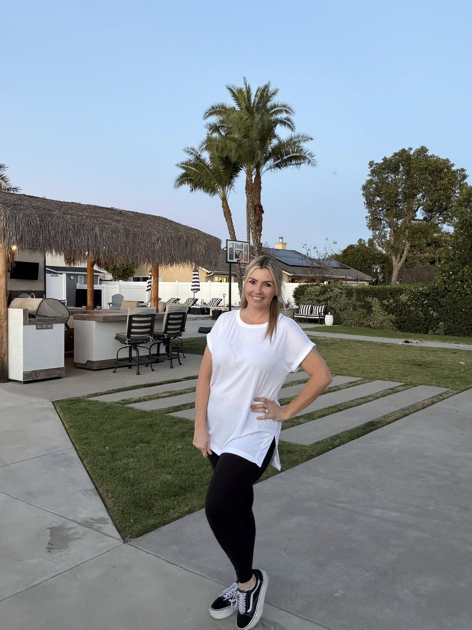 LuLaRoe Leah top in solid white, relaxed fit women’s white boutique top with soft drape fabric, classic everyday layering essential on a woman standing in a backyard with outdoor furniture and palm trees.