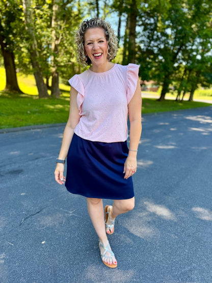 Woman in a LuLaRoe LeeAnn Ruffle Sleeve Top in Solid Blush Pink featuring floral eyelet fabric, double ruffle sleeves, crew neckline, relaxed fit, and a soft blush pink color and navy skirt standing on a paved path with trees in the background