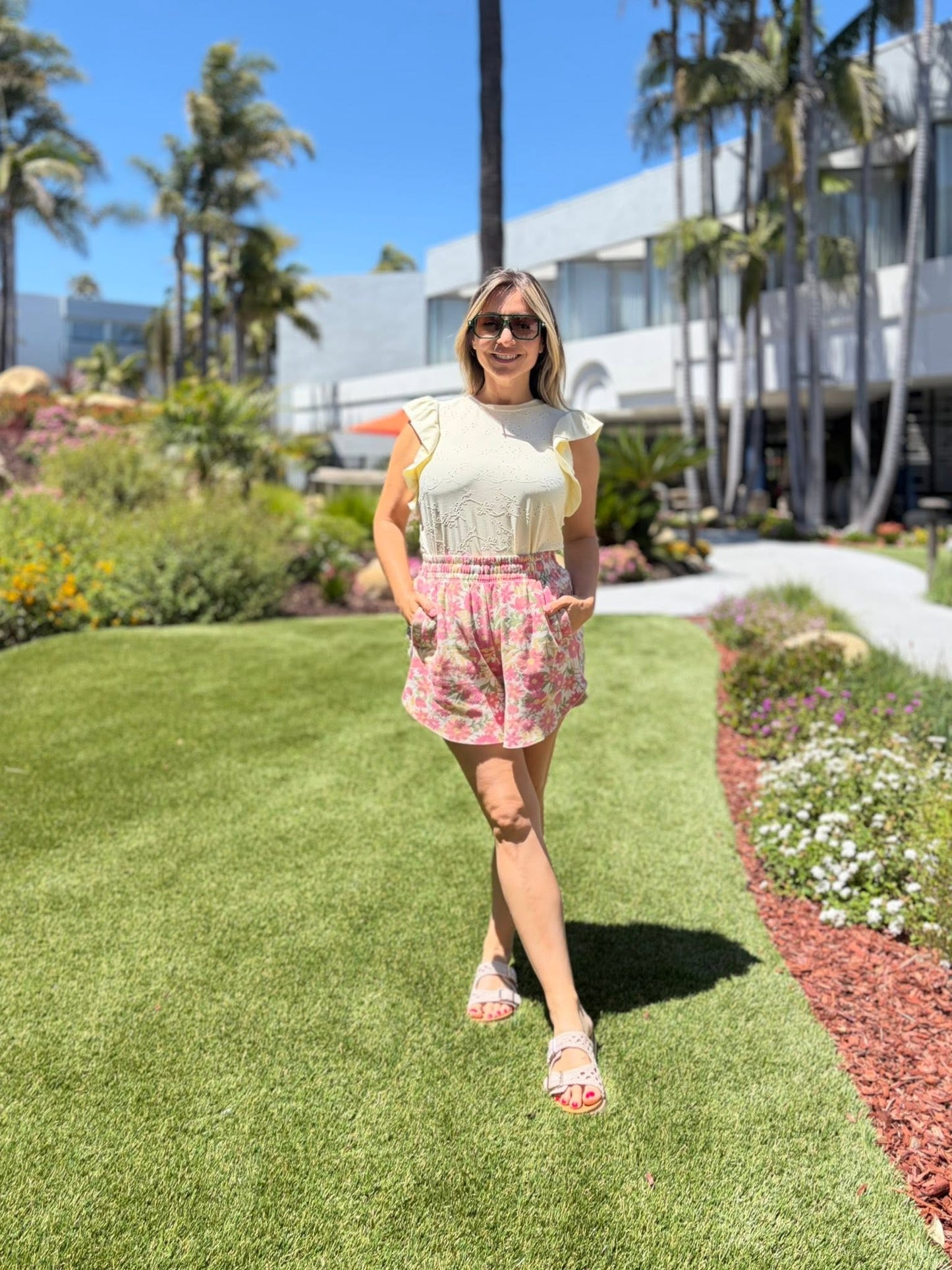 Woman wearing a LuLaRoe LeeAnn Ruffle Sleeve Top in Solid Buttercream featuring textured floral eyelet fabric, double ruffle sleeves, crew neckline, relaxed fit, and soft pastel yellow styling standing on grass with palm trees and a building in the background