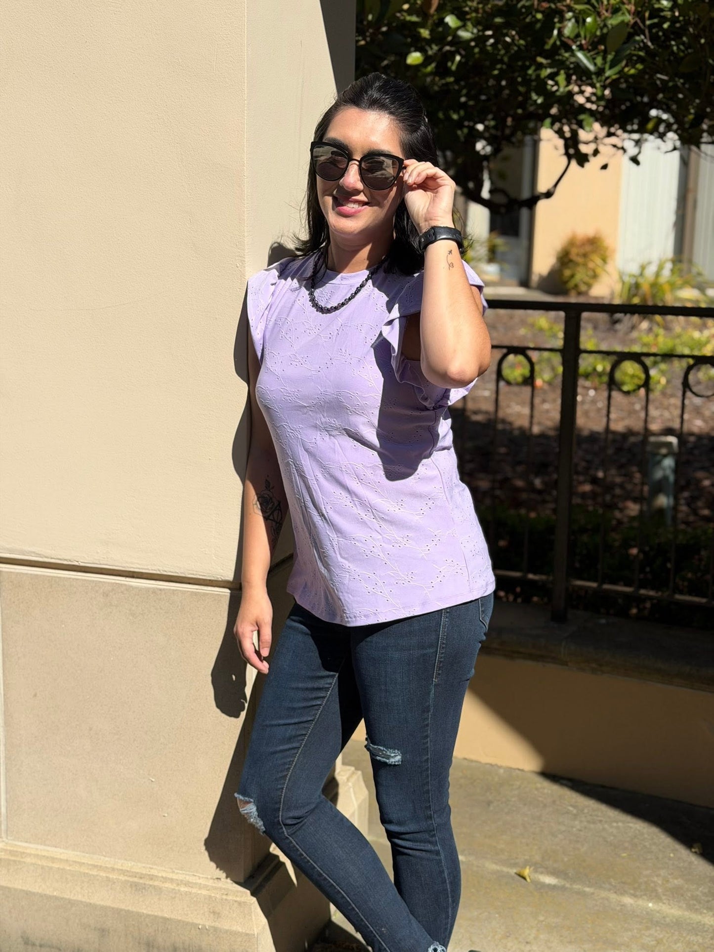 Woman wearing LuLaRoe LeeAnn Ruffle Sleeve Top in Solid Lavender Mist featuring floral eyelet fabric, double ruffle sleeves, crew neckline, relaxed fit, and a soft pastel lavender color and dark jeans standing outdoors.