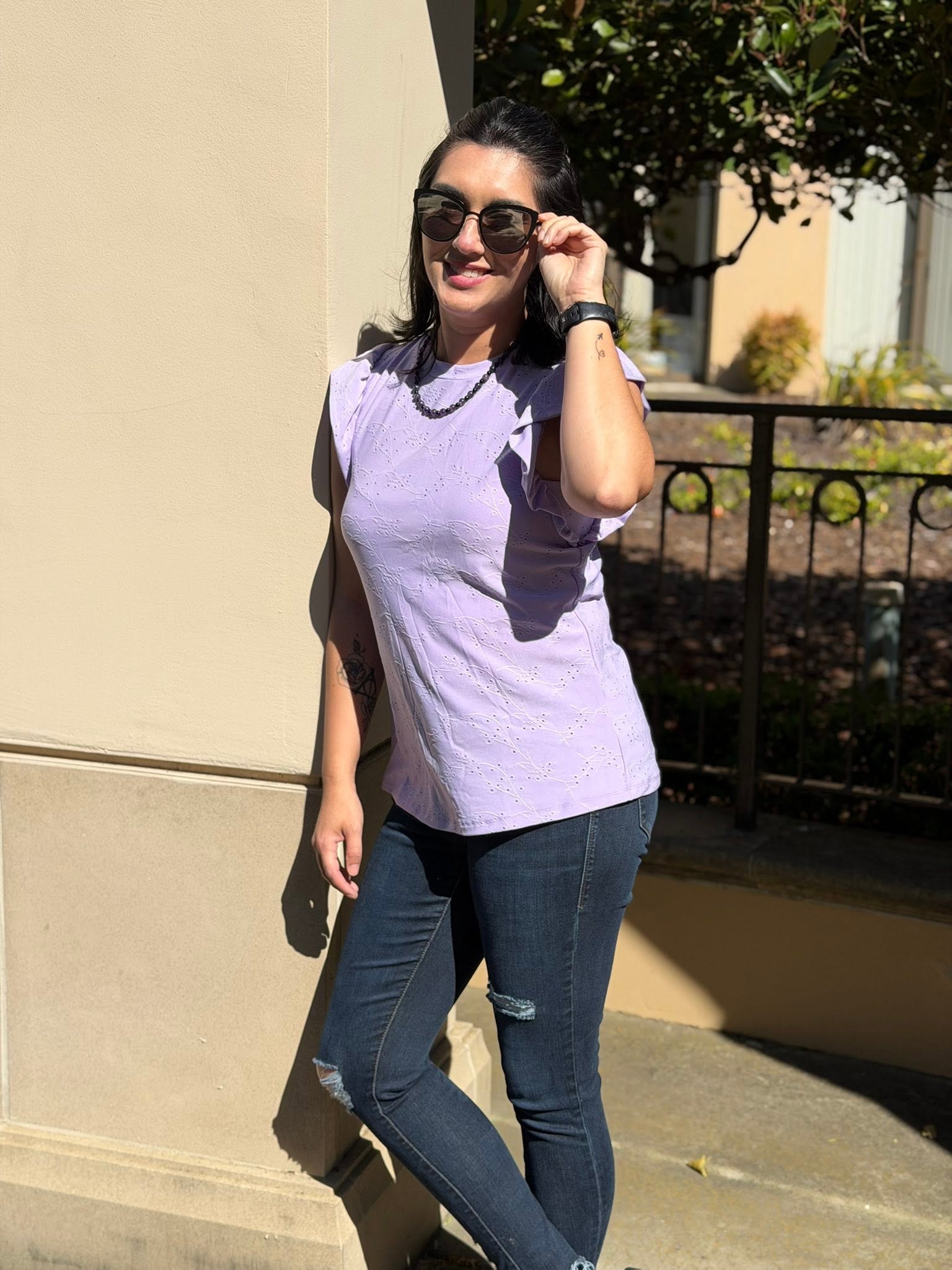 Woman wearing LuLaRoe LeeAnn Ruffle Sleeve Top in Solid Lavender Mist featuring floral eyelet fabric, double ruffle sleeves, crew neckline, relaxed fit, and a soft pastel lavender color and dark jeans standing outdoors.