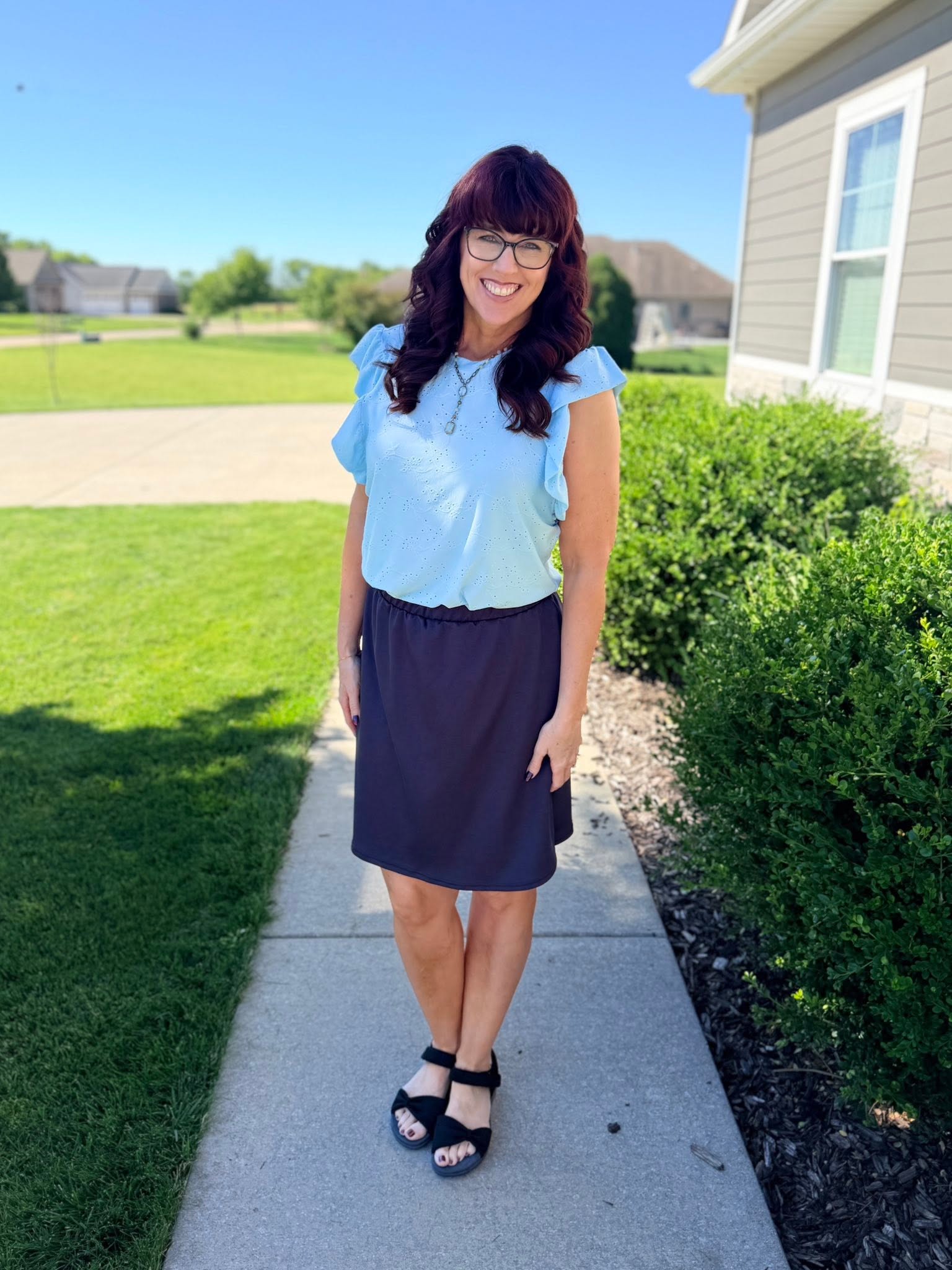 Woman in a LuLaRoe LeeAnn Ruffle Sleeve Top in Solid Sky Blue featuring textured floral eyelet fabric, double ruffle sleeves, crew neckline, relaxed fit, and a soft pastel blue color and navy skirt standing on a sidewalk with grass and a house in the background.