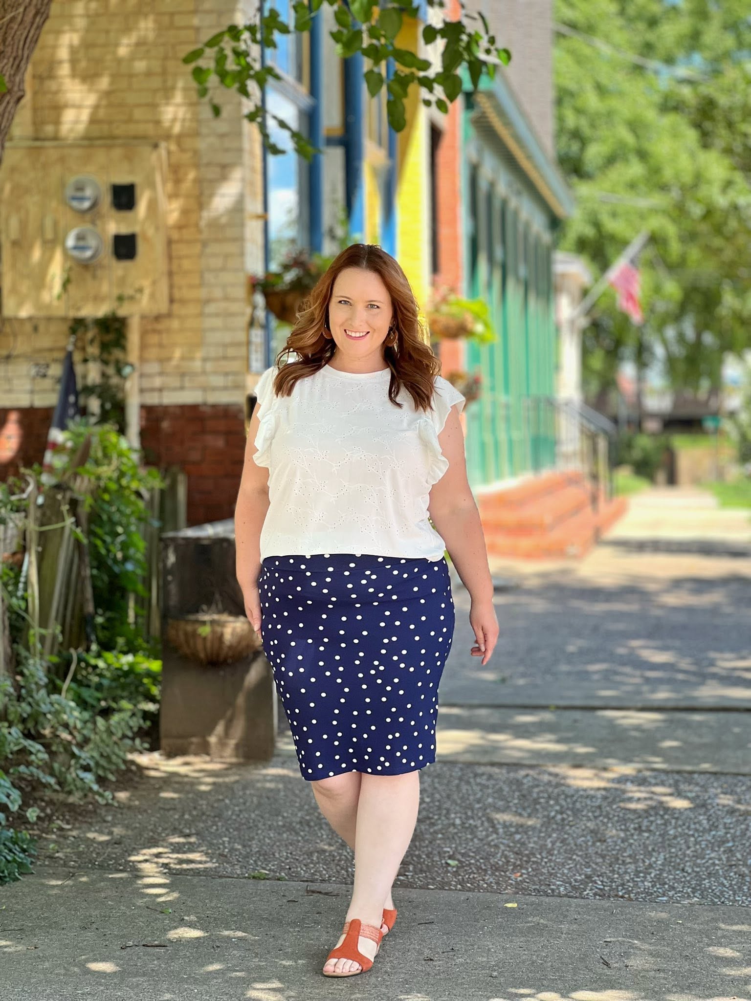 Woman in a LuLaRoe LeeAnn Ruffle Sleeve Top in Solid White featuring textured floral eyelet fabric, double ruffle sleeves, crew neckline, relaxed fit, and a timeless white color and navy skirt walking on a sidewalk with buildings and greenery in the background.
