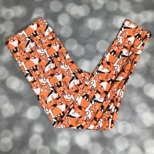 LuLaRoe leggings featuring an allover pattern of illustrated orange and tan corgi dogs on a white background, laid flat with crossed legs. on a blurred gray background