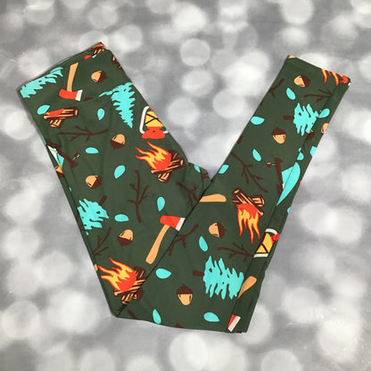 LuLaRoe leggings featuring a green background with illustrated campfire, axe, tree, acorn, and forest-themed icons in an all-over novelty print on a blurred natural background