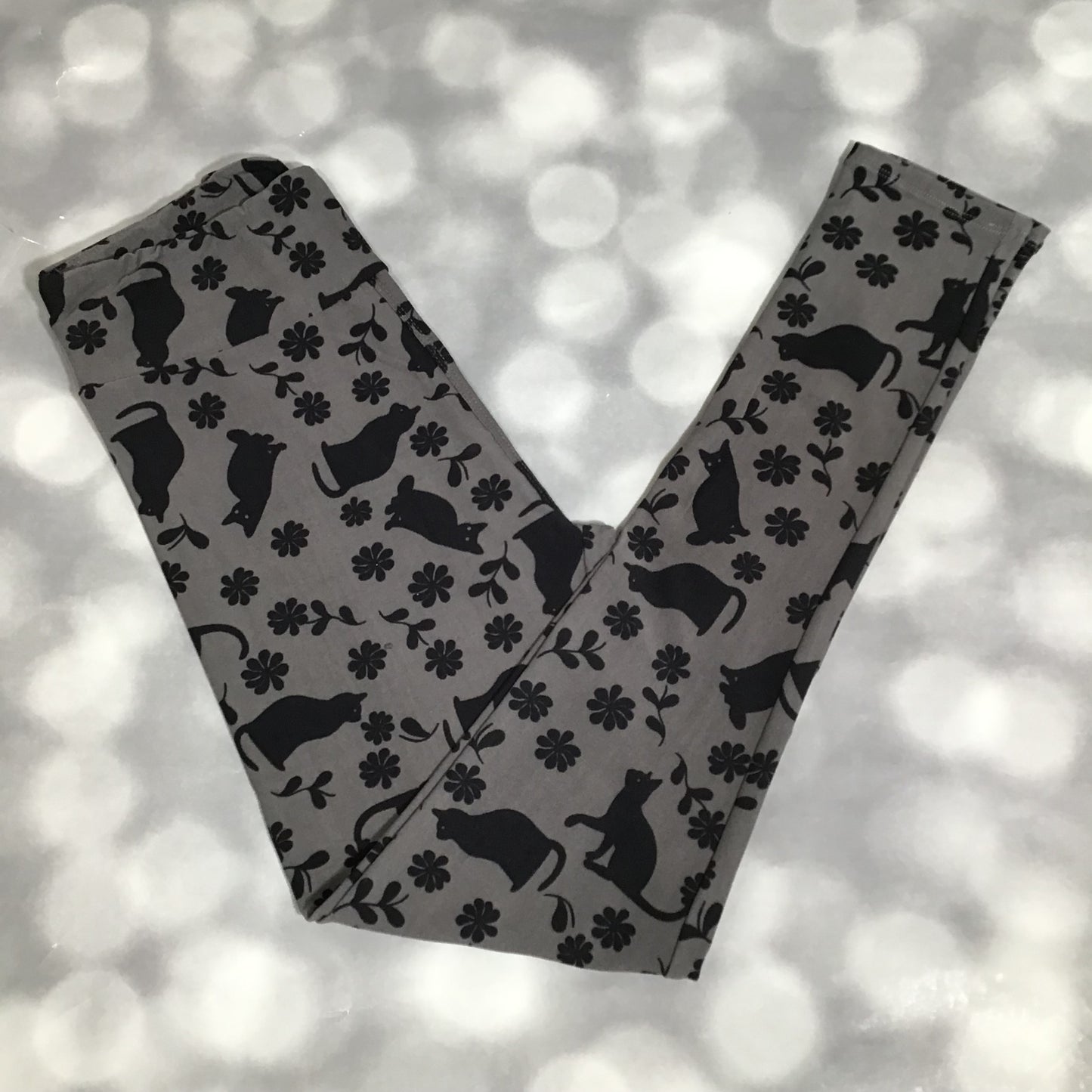 LuLaRoe leggings featuring a gray background with black silhouette cats, floral accents, and vine details in an all-over novelty pattern on a blurred natural background