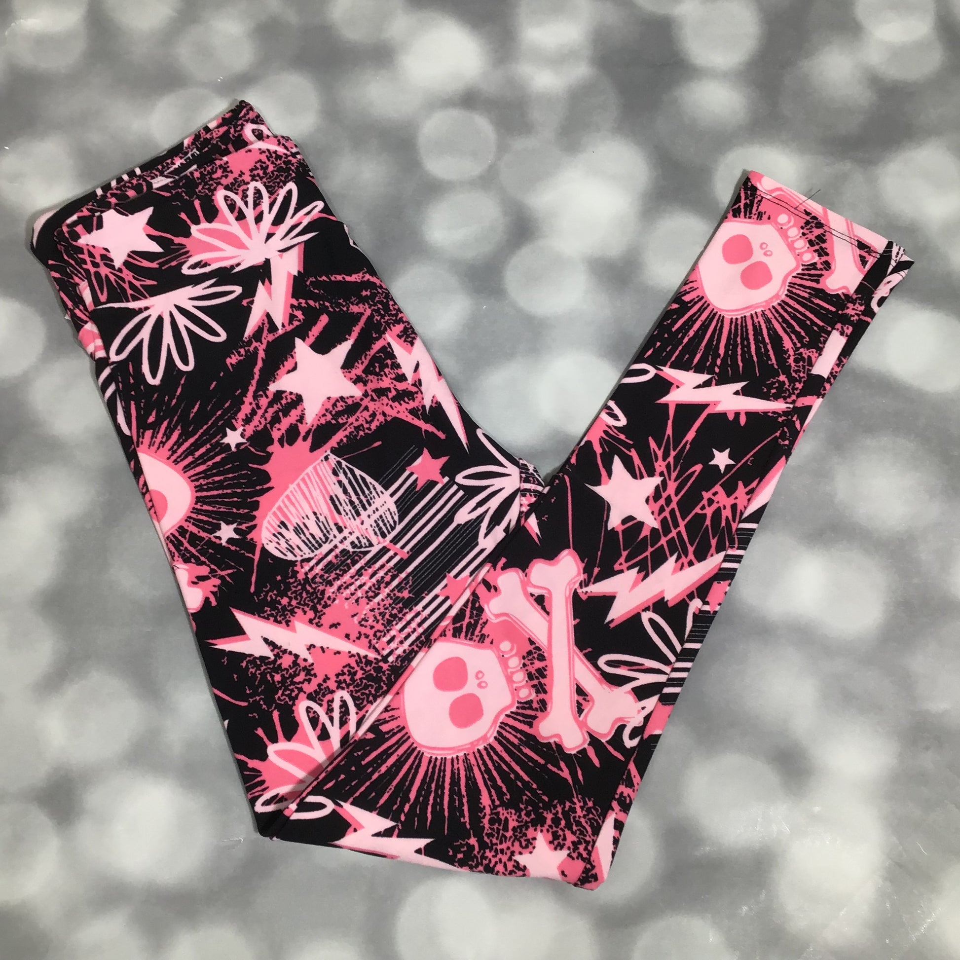 LuLaRoe leggings featuring a black background with neon pink skulls, bones, lightning bolts, and star graphics in an all-over edgy novelty print on a gray blurred background