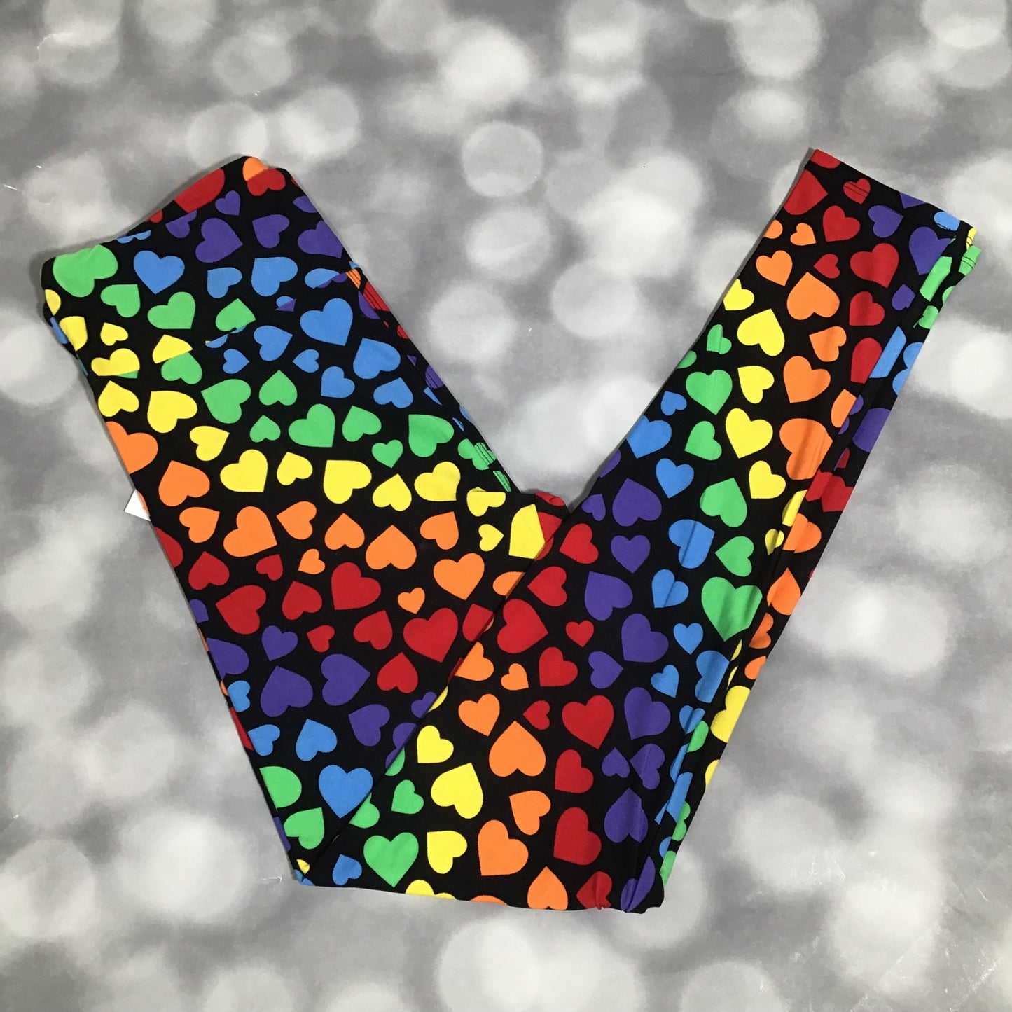 LuLaRoe leggings featuring a black background with multicolored rainbow heart shapes arranged in an all-over repeating pattern on a blurred gray background