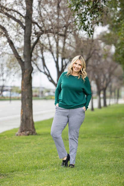 Woman modeling LuLaRoe Lena Work-Leisure Pants in Classic Noir Micro Houndstooth featuring a black and white small-scale houndstooth pattern with ankle length straight leg fit styled with a green sweater and black flats standing outdoors on grass with trees in the background
