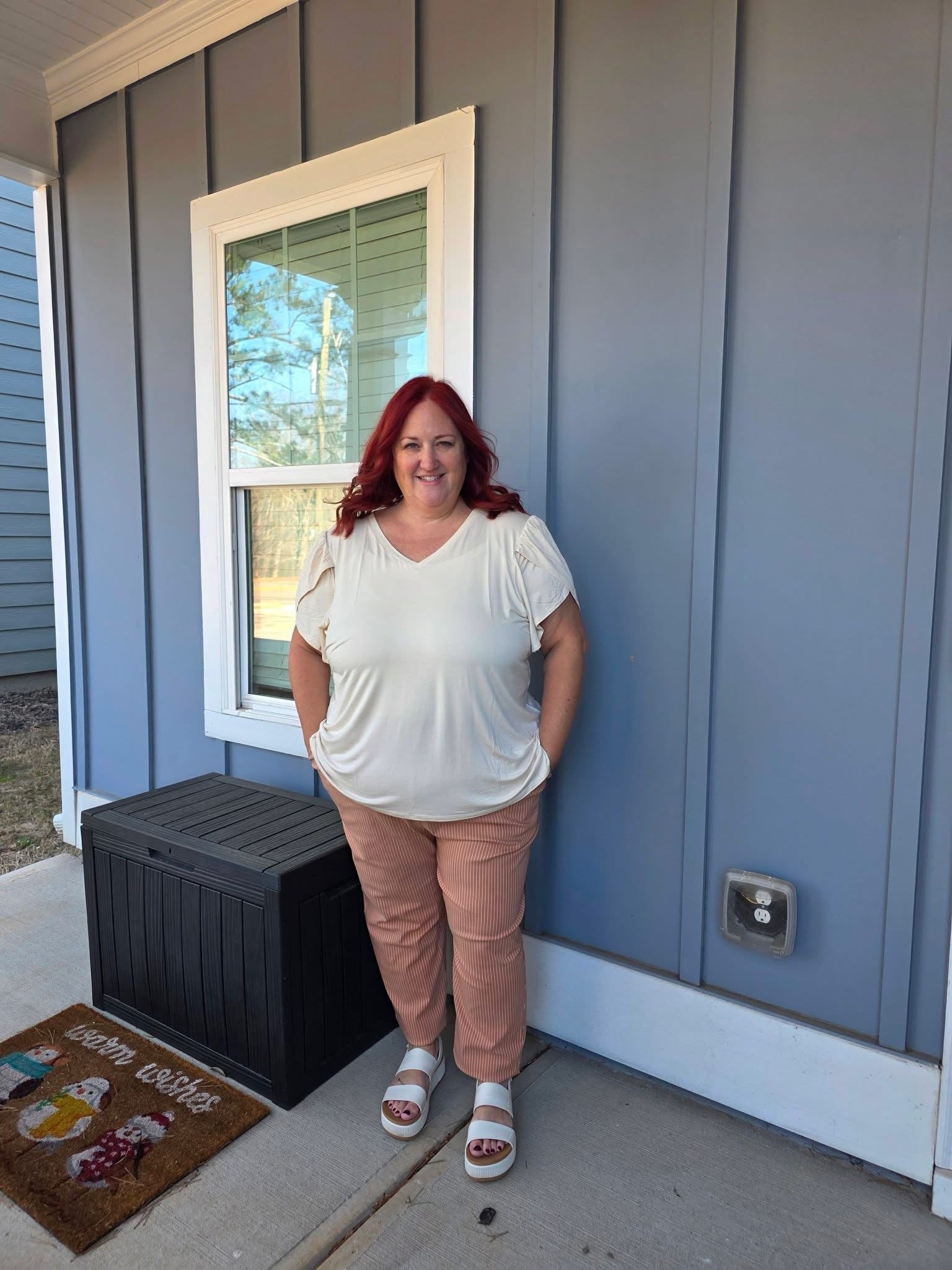 Woman modeling LuLaRoe Lena Work-Leisure Pants in Desert Clay Pinstripe featuring warm clay pink vertical pinstripe fabric with ankle length straight leg fit styled with a cream blouse and neutral sandals standing outside a house with a blue exterior