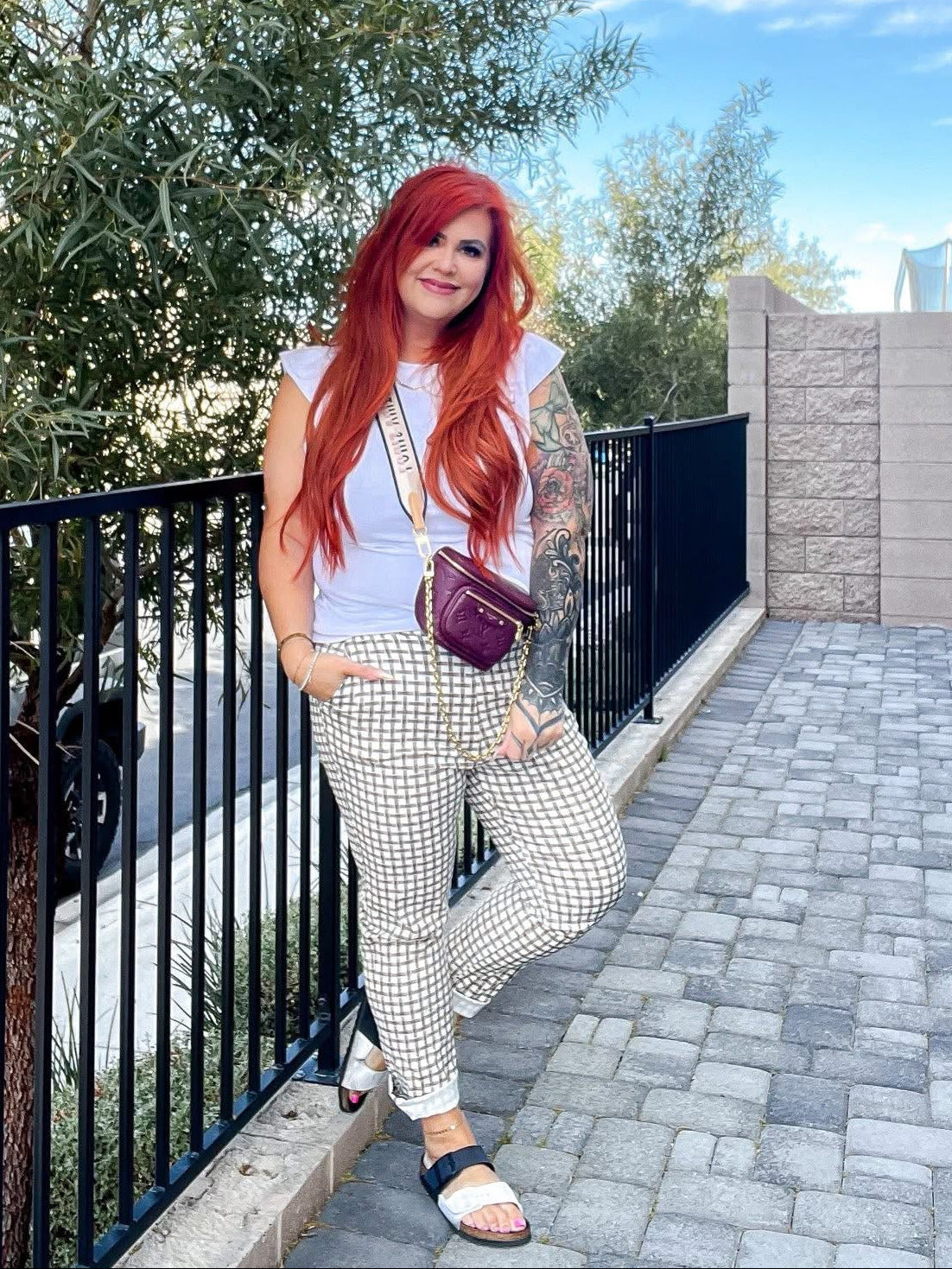 Woman modeling LuLaRoe Lena Work-Leisure Pants in Heritage Stone Gingham featuring a beige and ivory gingham check pattern with ankle length straight leg fit styled with a white sleeveless top and sandals standing outdoors with trees and buildings in the background