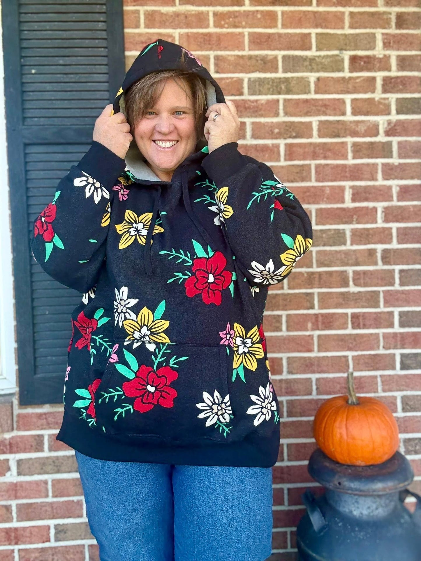 Woman wearing a black floral LuLaRoe Lexi Hooded Fleece Sweatshirt with bright red, yellow, and white flowers, featuring side slits, raglan sleeves, and a kangaroo pocket.