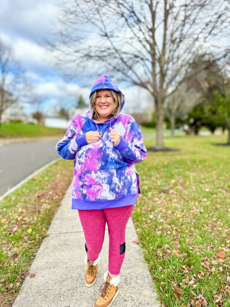 Woman wearing a purple and pink tie-dye LuLaRoe Lexi Hooded Fleece Sweatshirt with side slits, raglan sleeves, and a kangaroo pocket, styled outdoors with pink leggings.