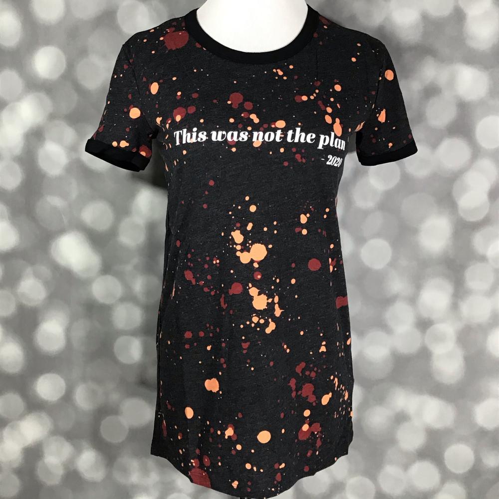 LuLaRoe Liv Tee featuring a black background with rust orange and burgundy paint splatter details and the phrase "This was not the plan - 2020" in white lettering. Women's graphic t-shirt with a humorous statement design and casual everyday style. on a gray background