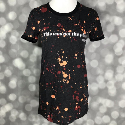LuLaRoe Liv Tee featuring a black background with rust orange and burgundy paint splatter details and the phrase "This was not the plan - 2020" in white lettering. Women's graphic t-shirt with a humorous statement design and casual everyday style. on a gray background