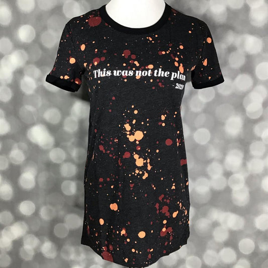 LuLaRoe Liv Tee featuring a black background with rust orange and burgundy paint splatter details and the phrase "This was not the plan - 2020" in white lettering. Women's graphic t-shirt with a humorous statement design and casual everyday style. on a gray background