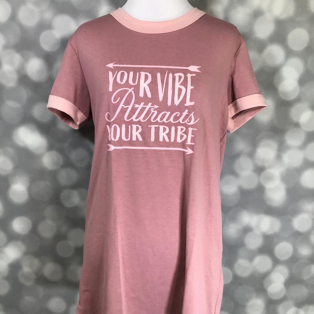 LuLaRoe Liv Tee in dusty rose pink featuring the inspirational phrase "Your Vibe Attracts Your Tribe" in blush pink lettering with arrow accents. Women's motivational graphic t-shirt with a positive message and casual everyday style on a blurred gray background