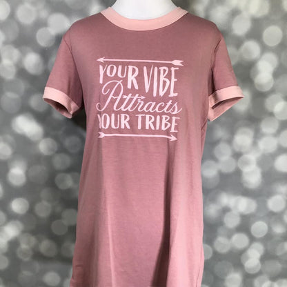 LuLaRoe Liv Tee in dusty rose pink featuring the inspirational phrase "Your Vibe Attracts Your Tribe" in blush pink lettering with arrow accents. Women's motivational graphic t-shirt with a positive message and casual everyday style on a blurred gray background
