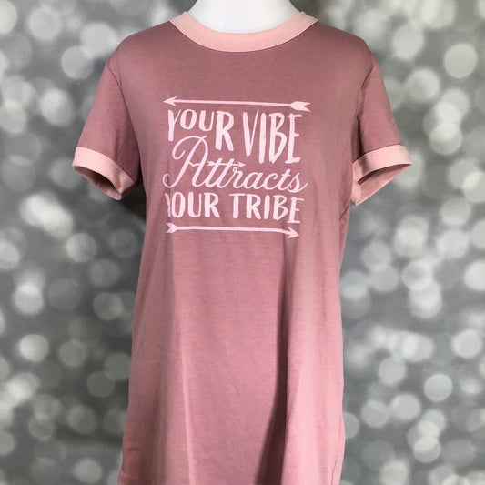 LuLaRoe Liv Tee in dusty rose pink featuring the inspirational phrase "Your Vibe Attracts Your Tribe" in blush pink lettering with arrow accents. Women's motivational graphic t-shirt with a positive message and casual everyday style on a blurred gray background