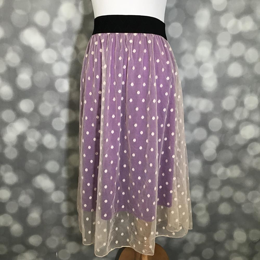 LuLaRoe Lola Skirt featuring a lavender purple layered mesh overlay with ivory swiss dot pattern and black elastic waistband displayed on a mannequin against a gray bokeh background. Feminine midi skirt with a flowy silhouette and romantic boutique-inspired styling.
