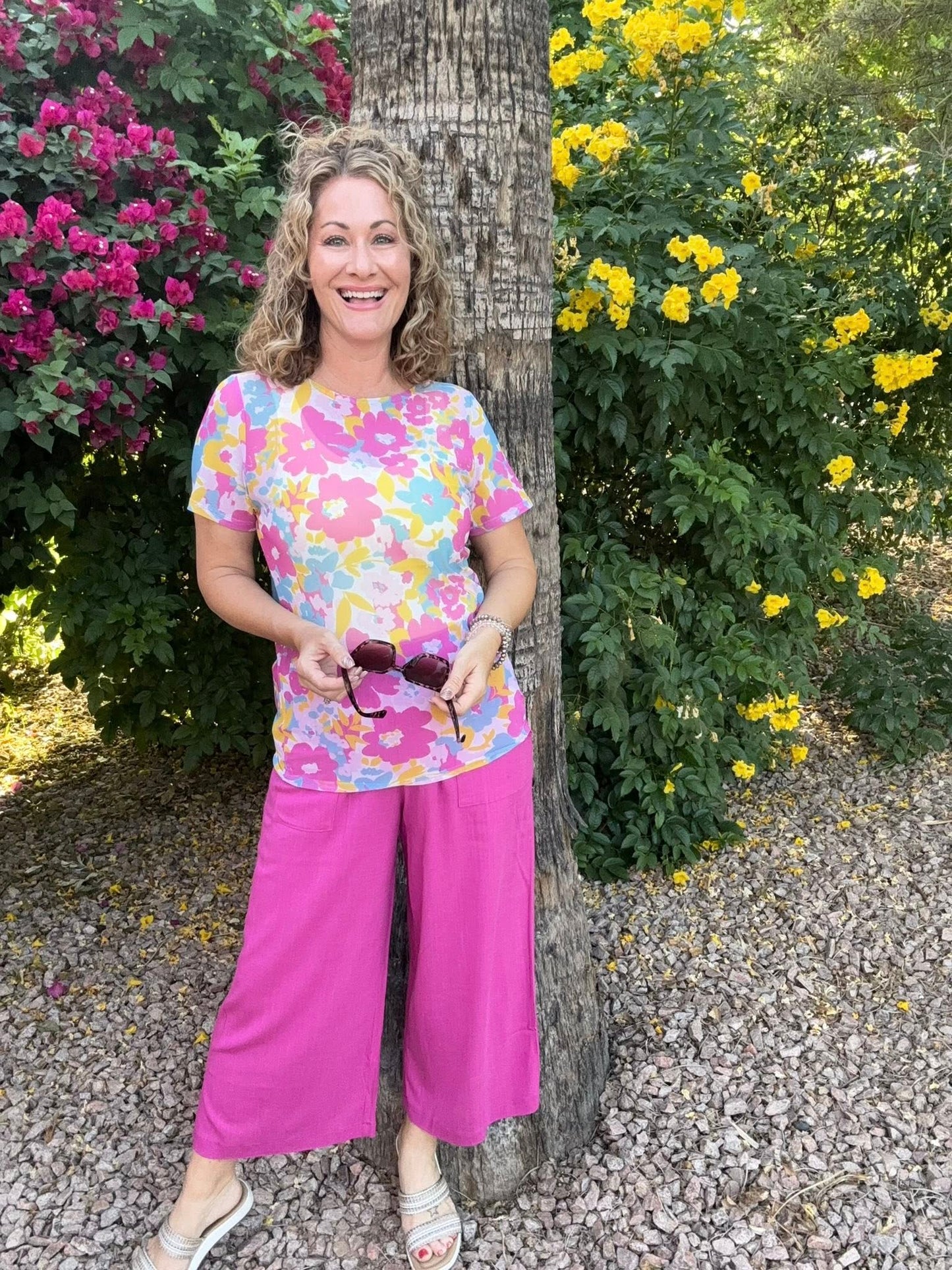Woman wearing a floral LuLaRoe Miranda Mesh T featuring bright pink, pastel blue, yellow, and lavender oversized flower prints paired with hot pink wide leg pants outdoors in a colorful tropical garden setting standing next to a tree with flowers in the background