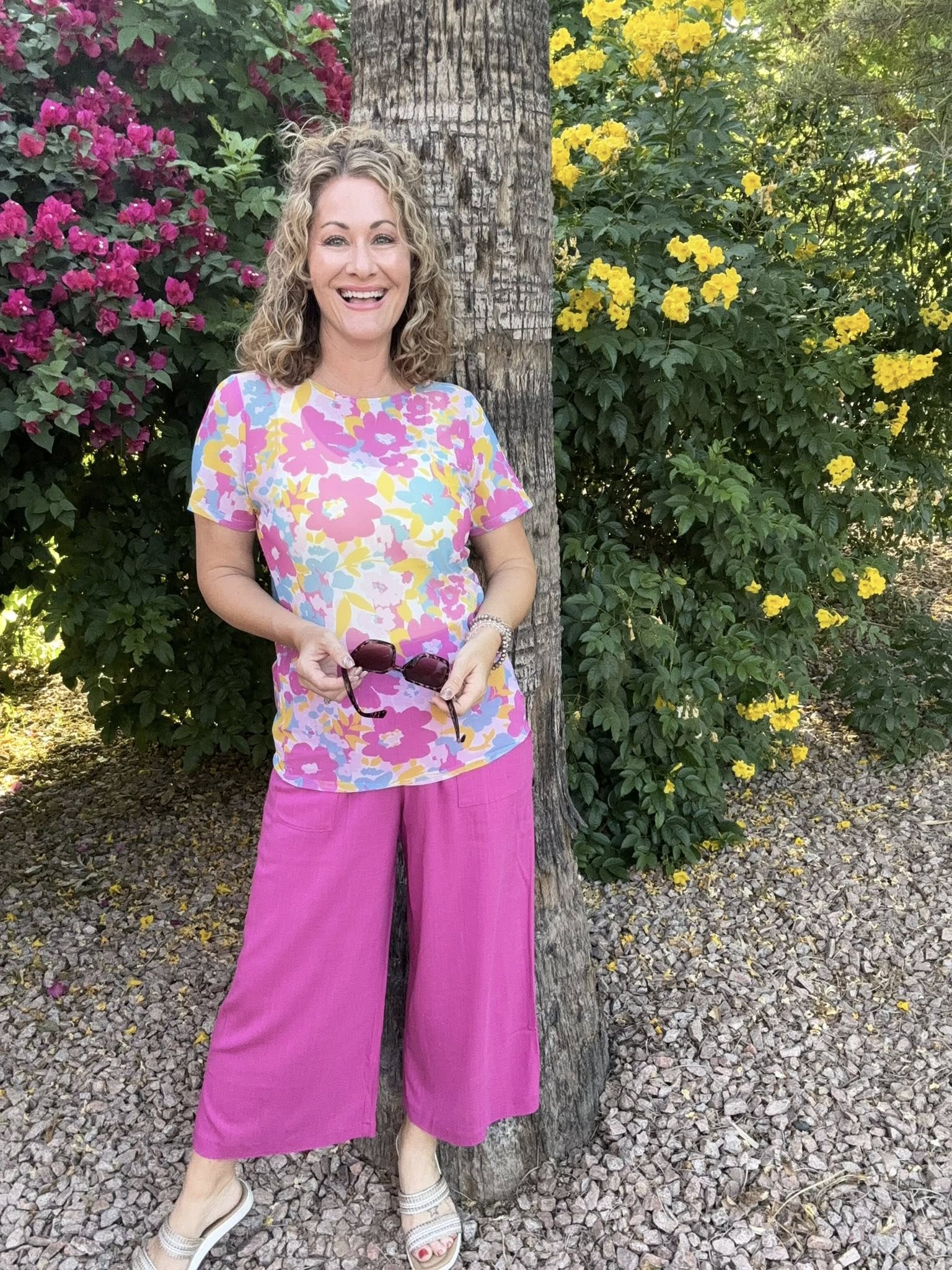 Woman wearing a floral LuLaRoe Miranda Mesh T featuring bright pink, pastel blue, yellow, and lavender oversized flower prints paired with hot pink wide leg pants outdoors in a colorful tropical garden setting standing next to a tree with flowers in the background