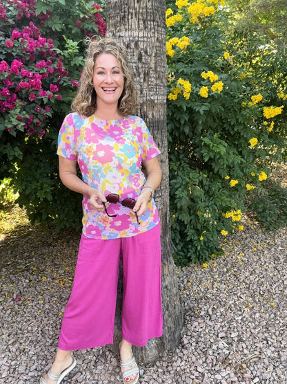 Woman wearing a floral LuLaRoe Miranda Mesh T featuring bright pink, pastel blue, yellow, and lavender oversized flower prints paired with hot pink wide leg pants outdoors in a colorful tropical garden setting standing next to a tree with flowers in the background