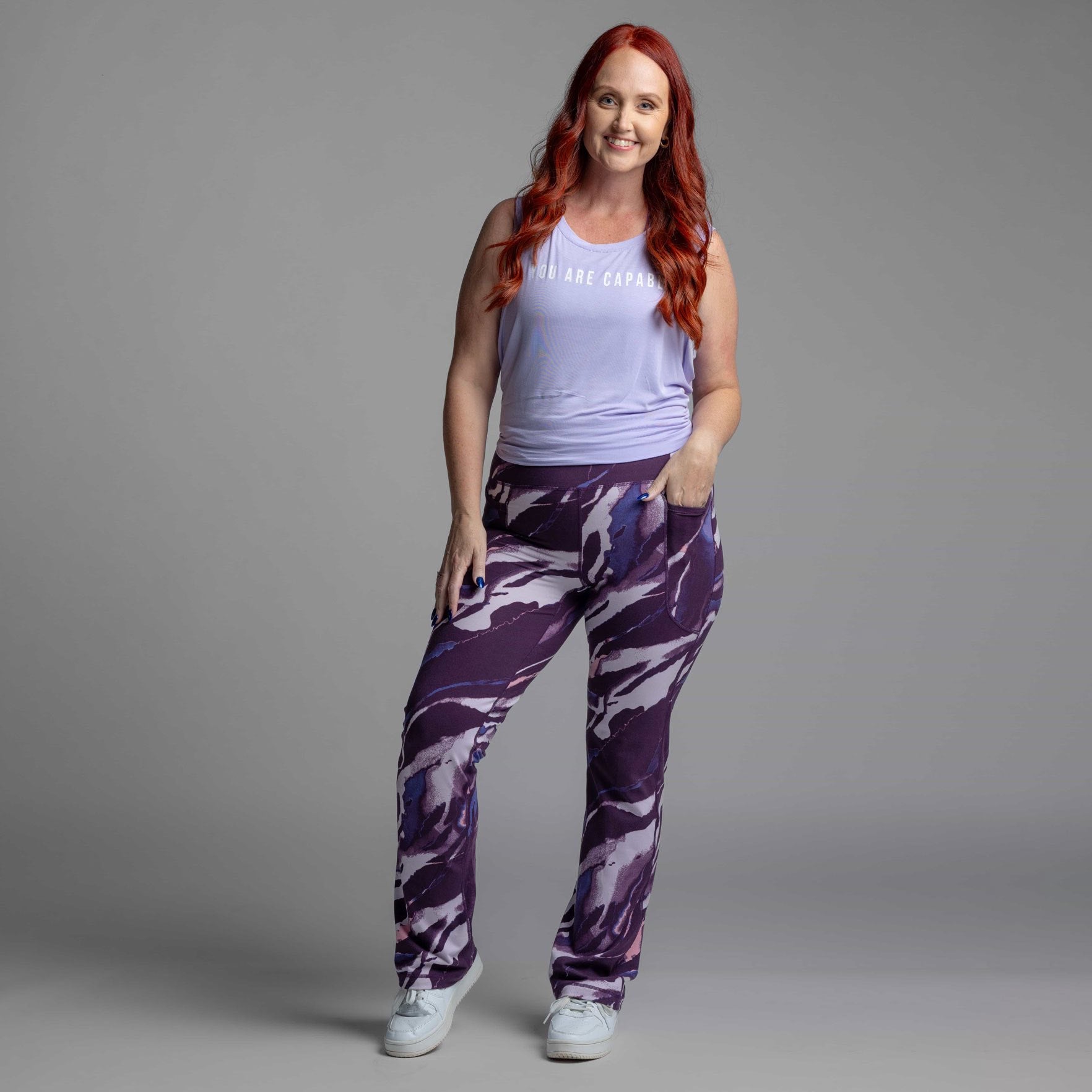 Woman wearing a light purple tank top and LuLaRoe Passionate Yoga Pant in amethyst vein abstract print with deep purple, charcoal, and soft lavender marbling, straight-leg fit, yoga waistband, and side hip pocket on a gray background