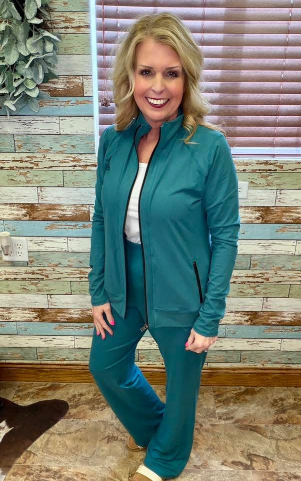 Woman wearing LuLaRoe Rise Bold jacket solid deep teal evergreen athletic zip jacket with black zipper and pockets standing in front of a wooden paneled wall.