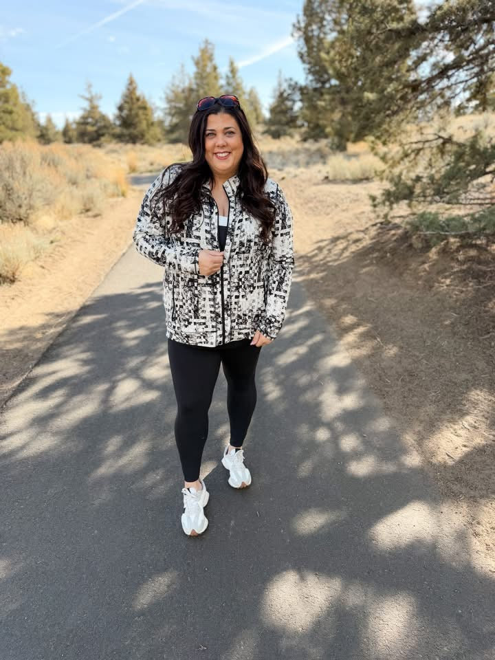 Woman wearing LuLaRoe Rise Bold jacket black and ivory pixel weave abstract athletic zip jacket with black zipper and pockets standing on a path with trees in the background