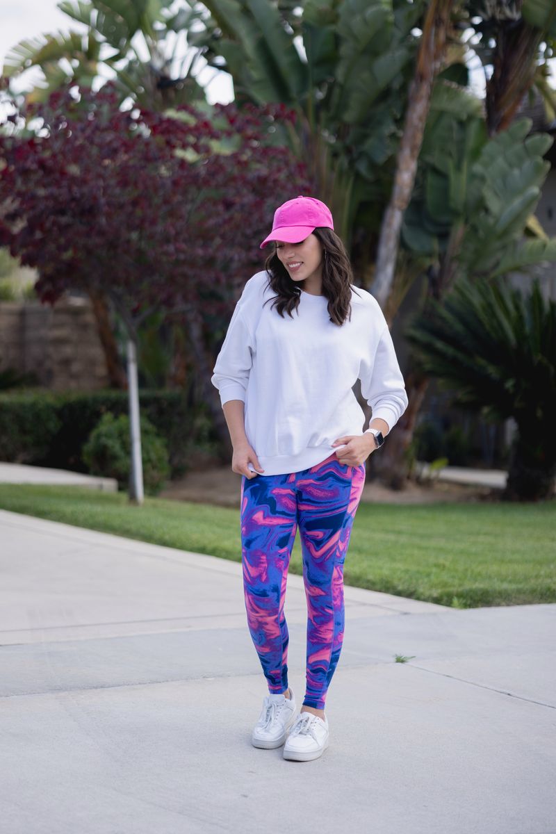 Woman wearing LuLaRoe Rise Brave leggings in pink and blue marble print styled with white sweatshirt and pink cap walking outdoors.