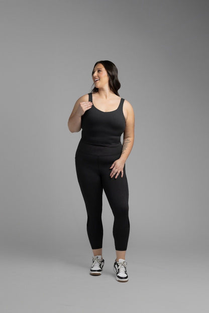 Woman modeling solid black LuLaRoe Rise Fearless Crop Leggings with a matching LuLaRoe Resilient Cami Bralette and white sneakers against a neutral gray studio background.