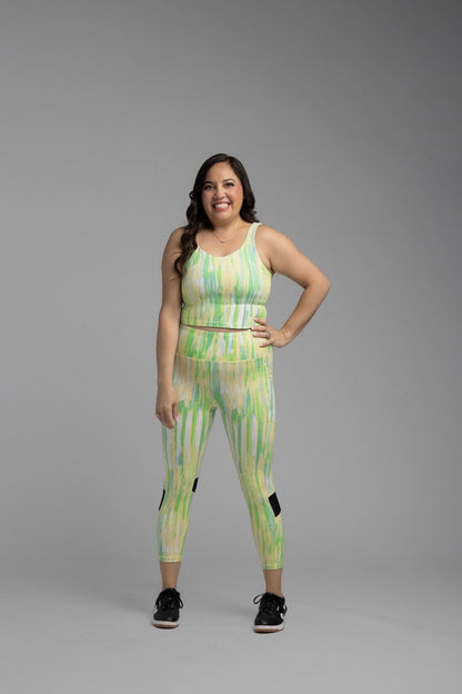 Woman modeling LuLaRoe Rise Fearless Crop Leggings in a pastel green and yellow watercolor abstract stripe print with black athletic sneakers against a neutral gray studio background.
