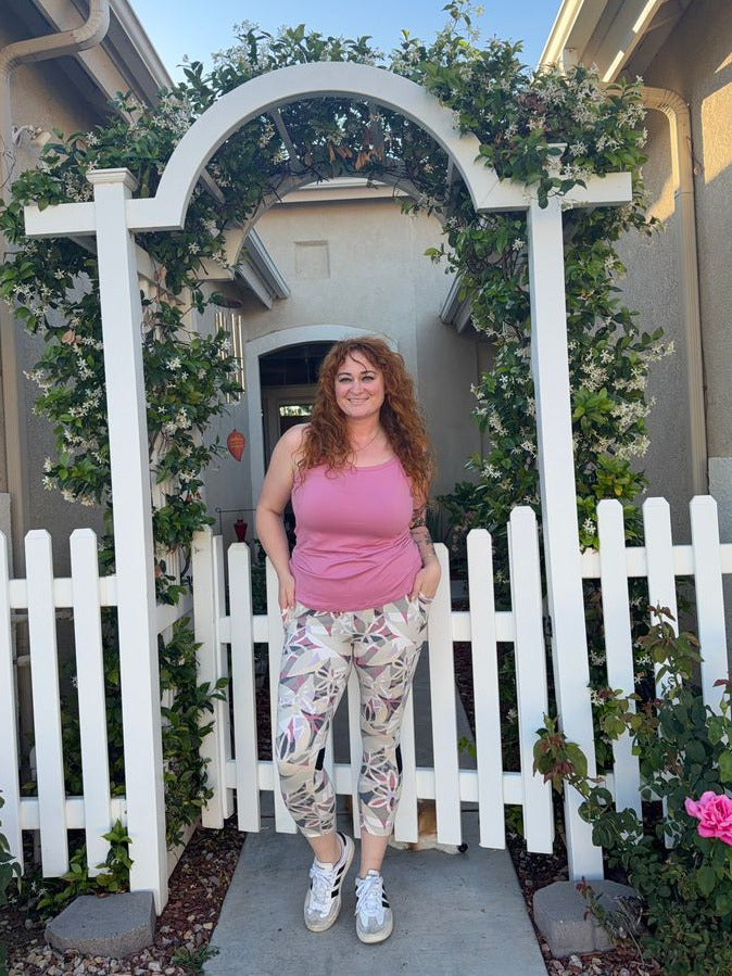 Woman modeling LuLaRoe Rise Fearless Crop Leggings in a blush pink, taupe, cream, and mauve abstract geometric floral print paired with a pink tank top and white sneakers outdoors near a garden archway.