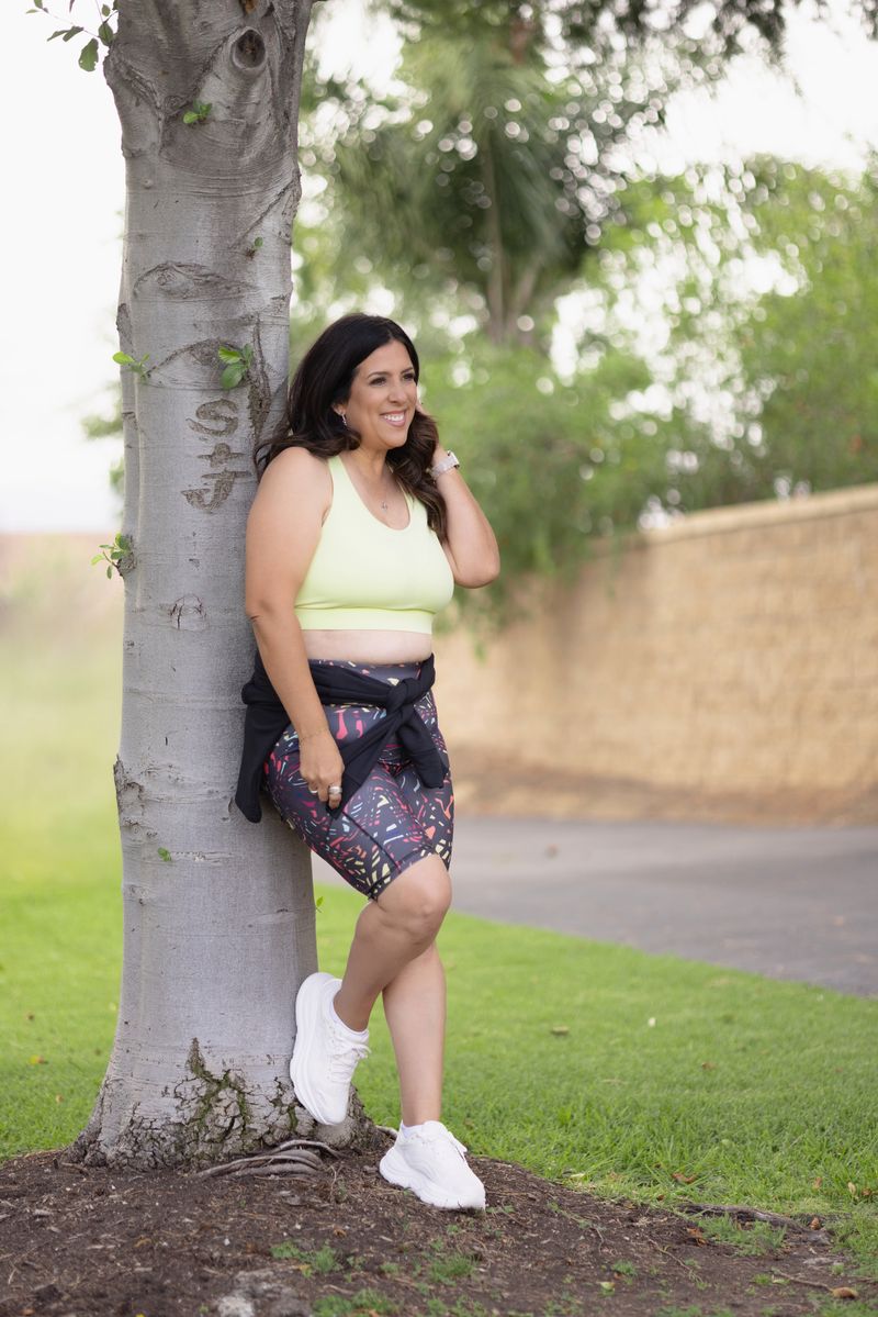 Woman leaning against a tree wearing a LuLaRoe RISE Invincible Sports Bra in solid butter yellow with a racerback design, paired with patterned biker shorts, styled for activewear comfort outdoors.