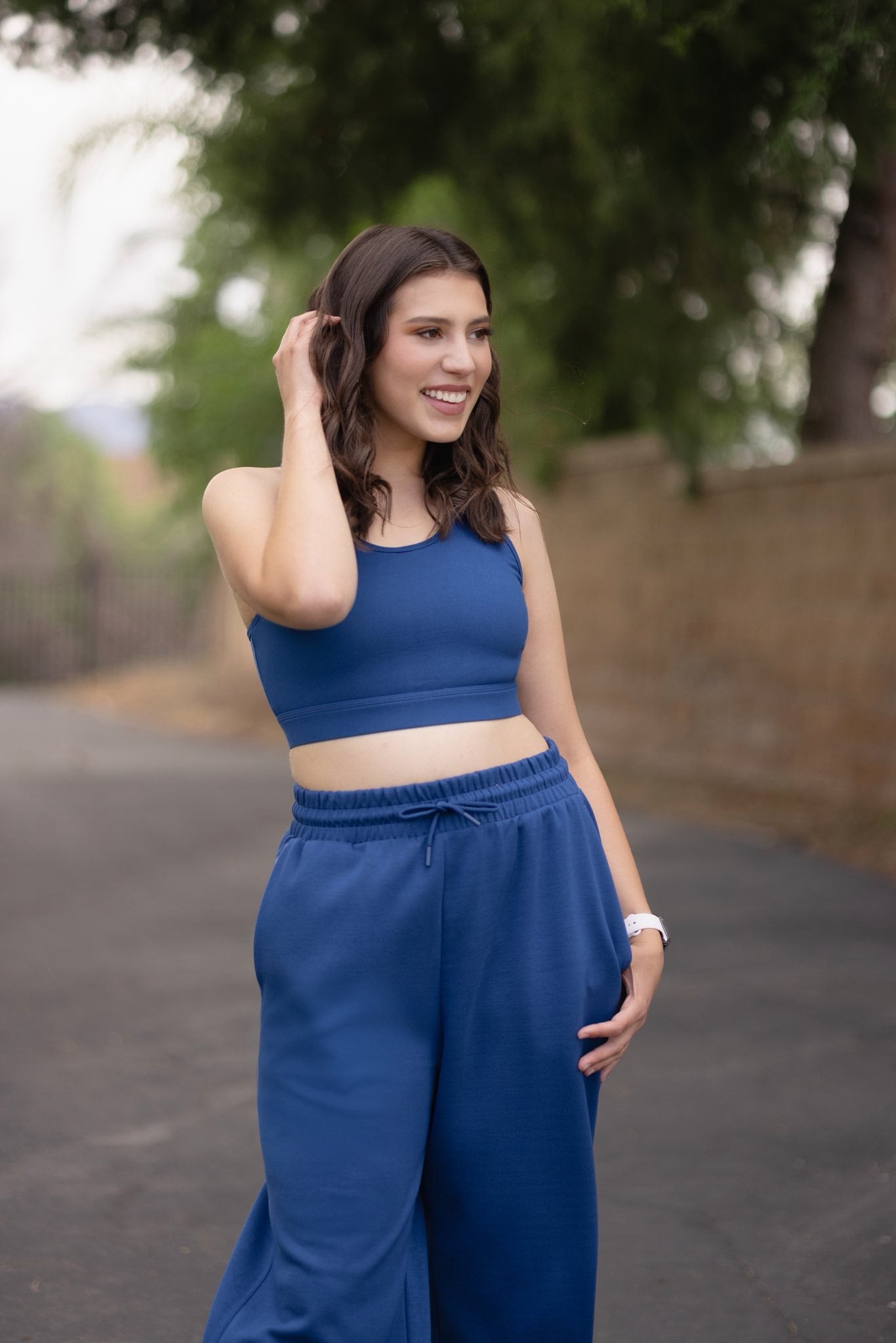 Woman wearing a solid peony blue LuLaRoe Rise Invincible sports bra with racerback silhouette and matching blue athleisure pants, styled outdoors with a clean, modern activewear look with trees in the background